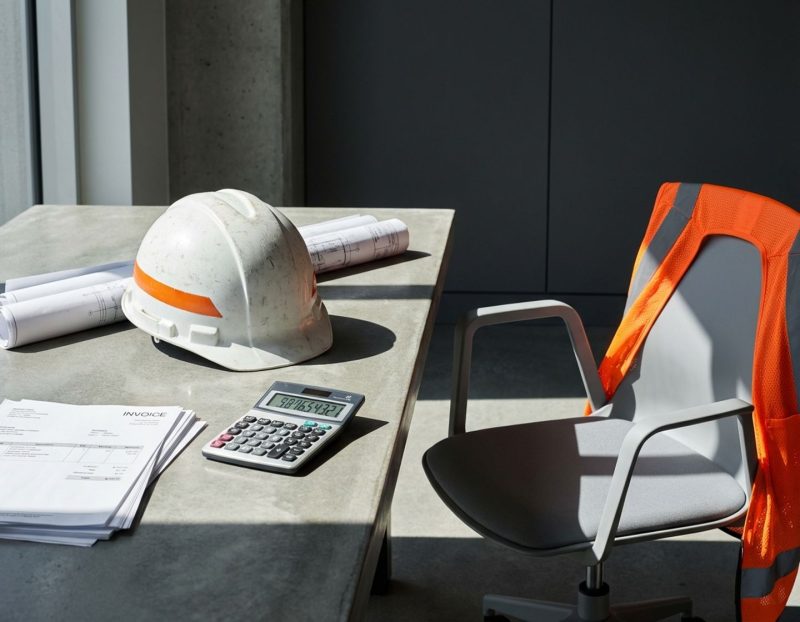lege bouwhelm en opgerolde blauwdrukken op betonnen bureau met rekenmachine, facturen en oranje veiligheidsvest