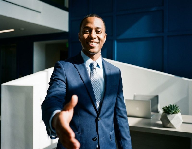 zelfverzekerde zakenman in marineblauw pak steekt hand uit voor handdruk in modern kantoor met laptop en plant